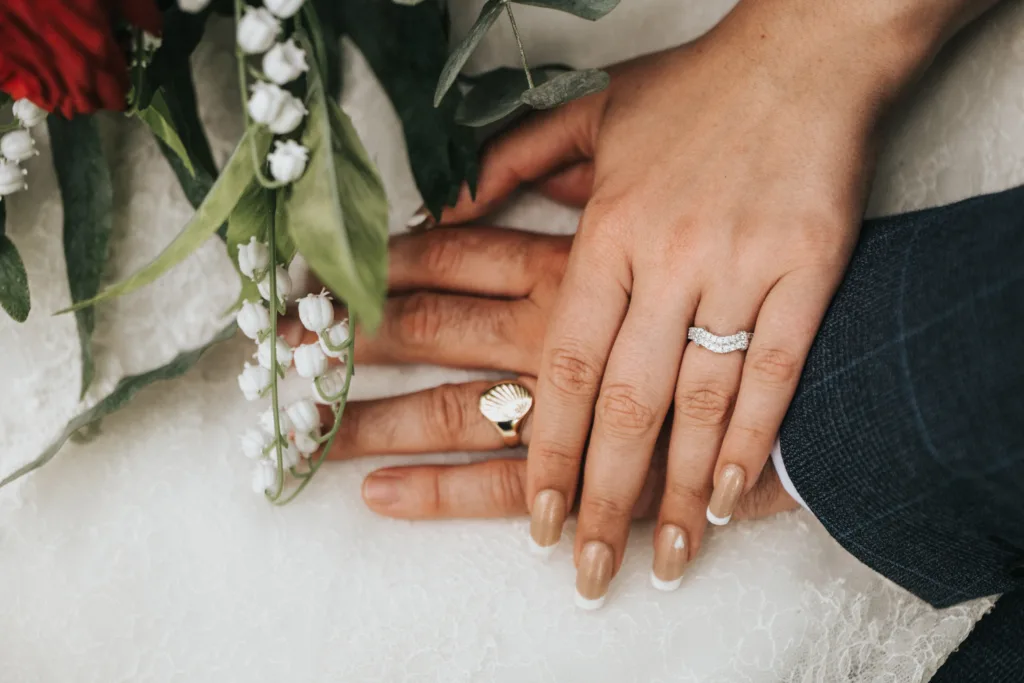 Two hands rest together on white lace, one with a diamond ring and French manicure, the other with a gold band. A bouquet of red and white flowers is nearby. © Aimee Lince Photography, wedding and lifestyle photographer in Lincolnshire