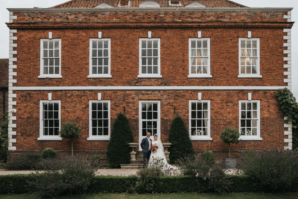 A Bardney Hall bride and groom stand together on the steps in front of a large, elegant brick building with white-trimmed windows, surrounded by greenery and neatly manicured bushes. © Aimee Lince Photography, wedding and lifestyle photographer in Lincolnshire