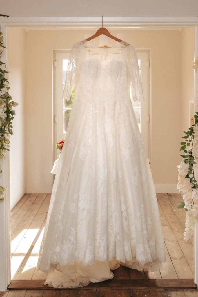 A white lace wedding dress with long sleeves and a full skirt hangs on a wooden hanger in a bright room with wooden floors and floral decorations on the doorframe, sunlight streaming through windows behind it. © Aimee Lince Photography, Lincolnshire photographer