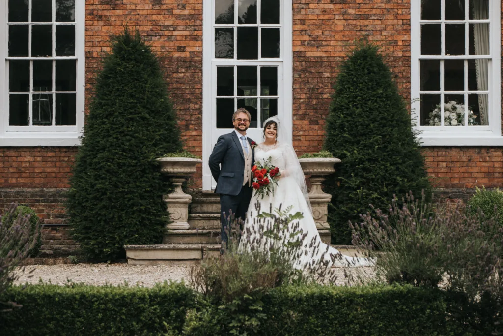A bride in a white dress and veil holding a red bouquet stands next to a groom in a blue suit and bow tie. They pose outside in front of a red brick building with tall windows, flanked by two large, cone-shaped evergreen shrubs. © Aimee Lince Photography, Lincolnshire photographer