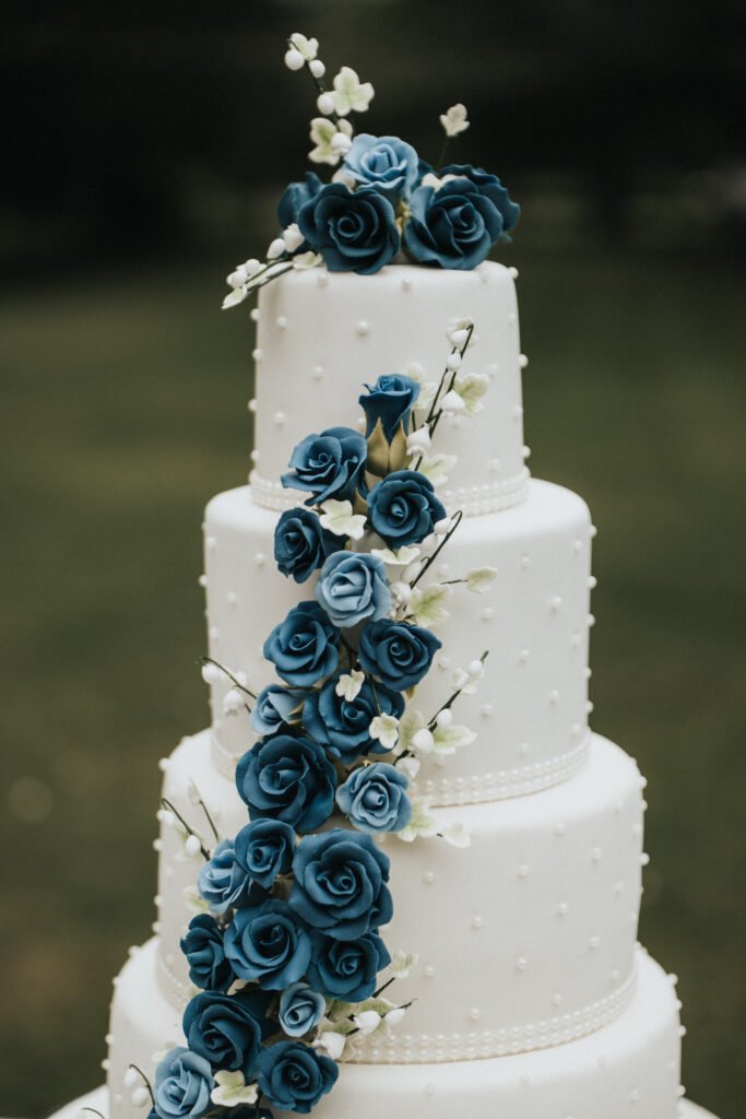 A four-tier white wedding cake decorated with small piped dots and adorned with cascading blue roses and small white flowers made of fondant or sugar. The cake is set outdoors on a blurred green background. © Aimee Lince Photography, Lincolnshire photographer