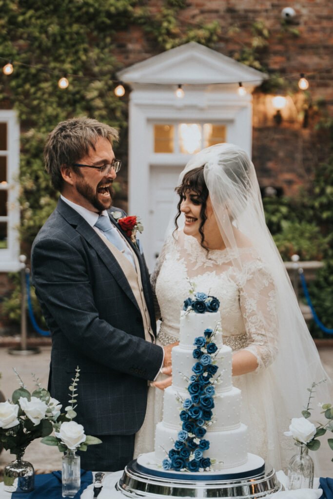 A bride in a lace wedding dress and veil stands beside a groom in a suit and tie. Both are smiling and cutting a white wedding cake decorated with cascading blue roses. White flowers in vases are on the table; a brick wall and door are in the background. © Aimee Lince Photography, Lincolnshire photographer