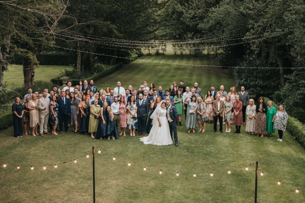 A large group of wedding guests stands on a green lawn, gathered around a bride in a white dress and a groom in a dark suit. Fairy lights hang overhead, and tall trees and hedges frame the grassy background. Everyone is smiling and dressed up. © Aimee Lince Photography, Lincolnshire photographer