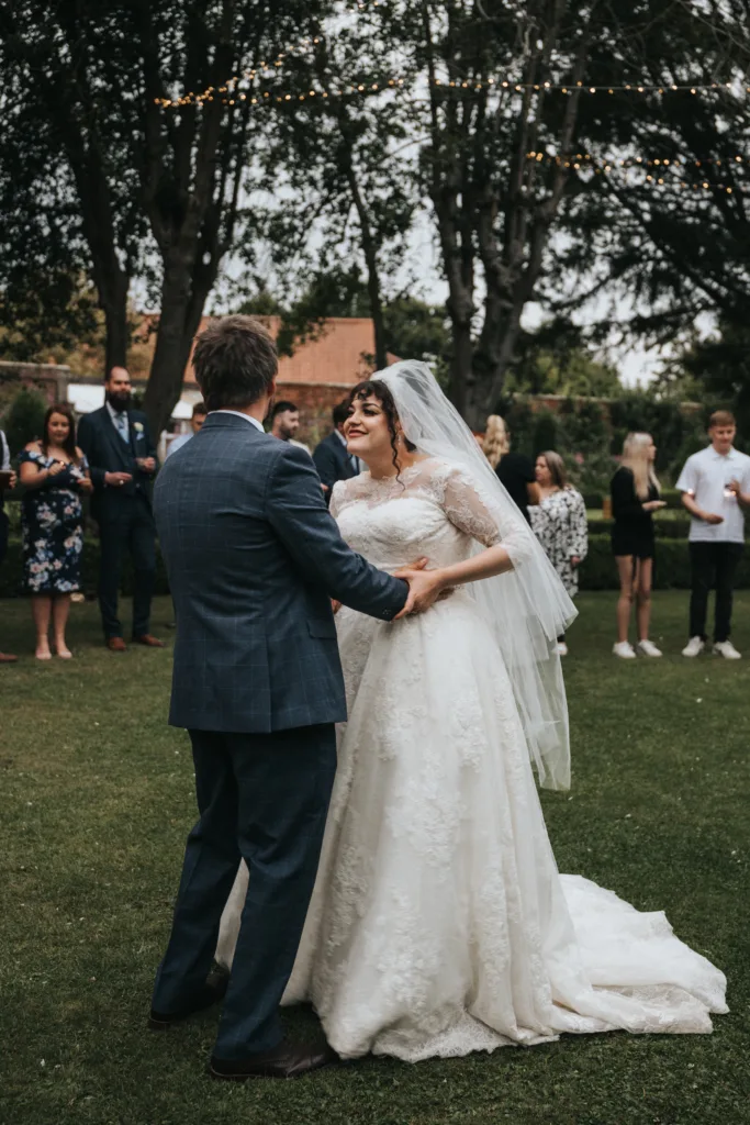 A bride in a lace wedding dress and veil smiles whilst dancing with the groom in a blue suit on grass. Guests stand and watch in the background, some talking and others holding drinks, under fairy lights and trees in an outdoor setting. © Aimee Lince Photography, Lincolnshire photographer