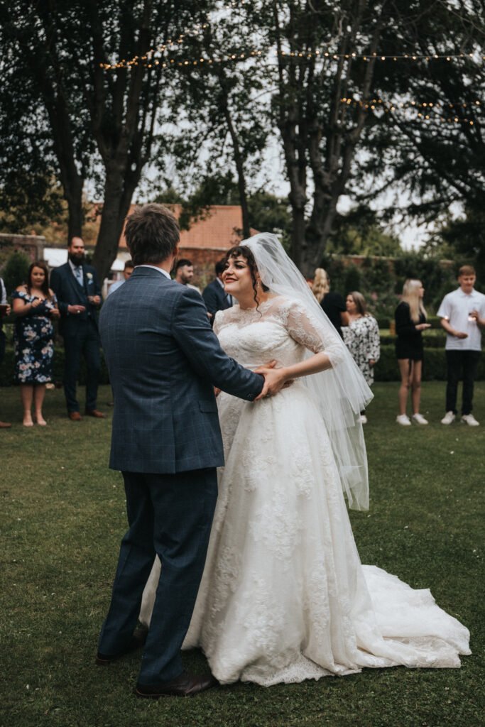 A bride in a lace wedding dress and veil smiles whilst dancing with the groom in a blue suit on grass. Guests stand and watch in the background, some talking and others holding drinks, under fairy lights and trees in an outdoor setting. © Aimee Lince Photography, Lincolnshire photographer