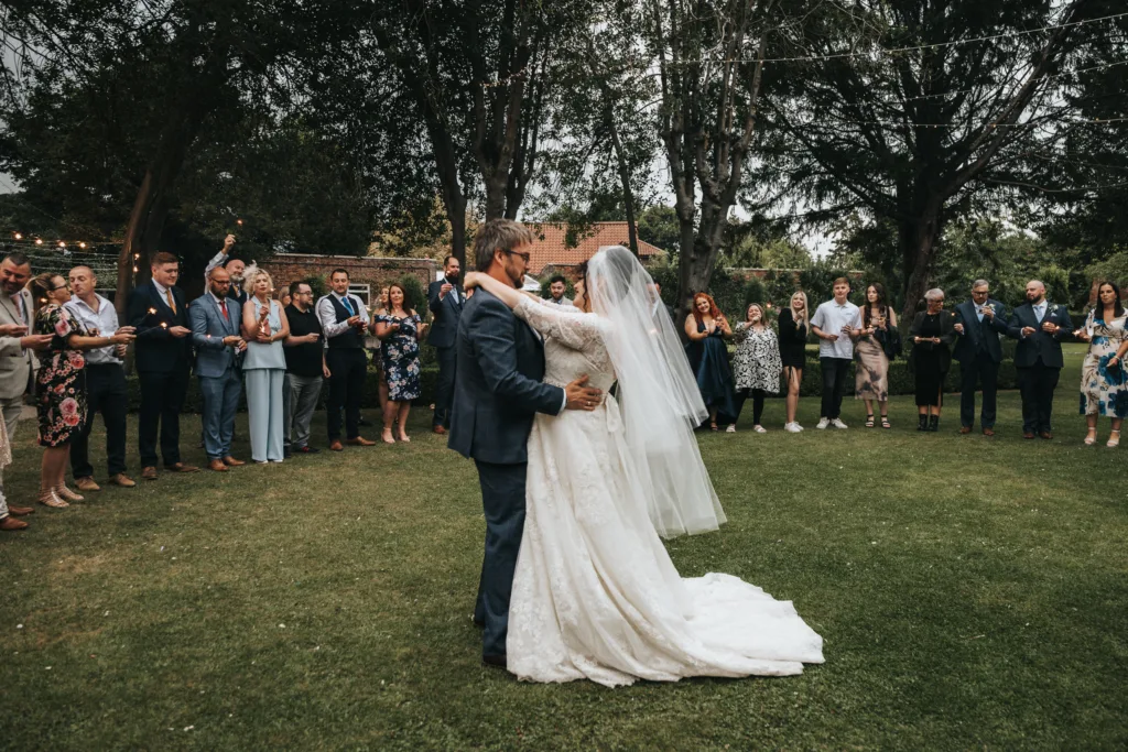 A bride in a white dress and veil dances with the groom in a blue suit on grass, surrounded by guests forming a circle outdoors. Trees and fairy lights are above, and people are smiling, clapping, and watching the couple’s first dance. © Aimee Lince Photography, Lincolnshire photographer