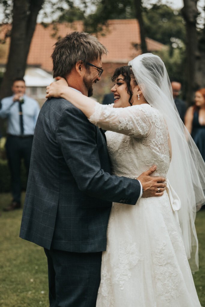 A bride in a lace wedding dress and veil smiles joyfully as she embraces a bearded groom in a checked suit. They are dancing outdoors on grass, surrounded by guests in formal attire, with blurred trees and buildings in the background. © Aimee Lince Photography, Lincolnshire photographer