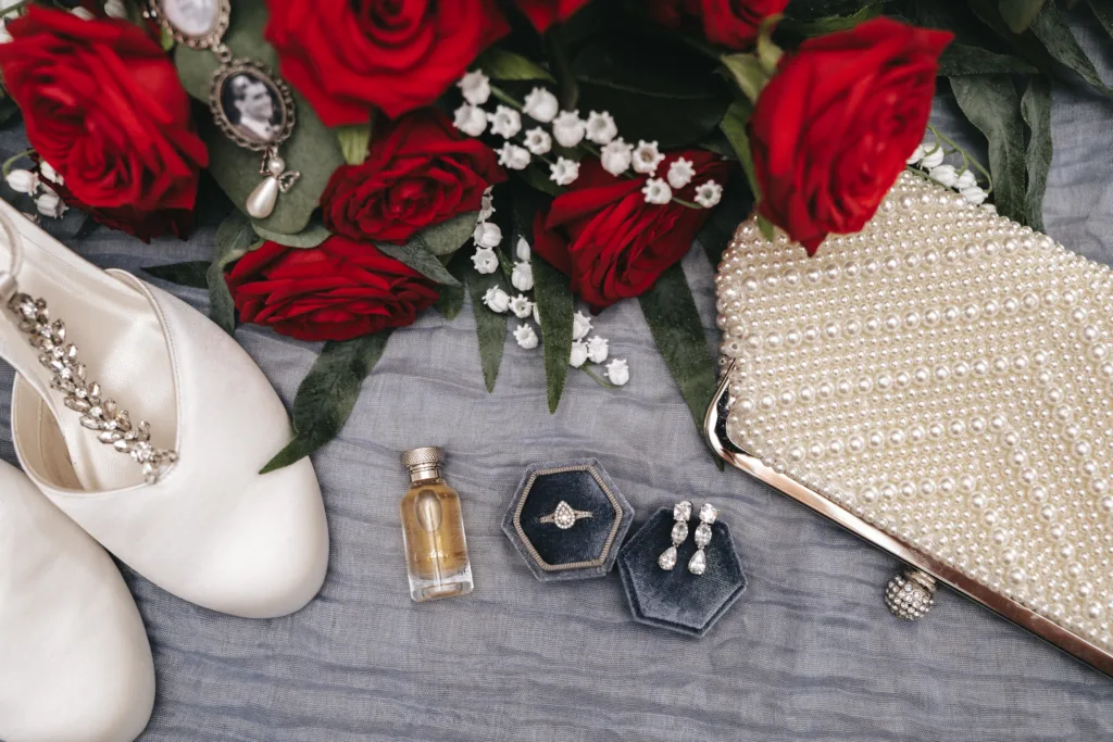 A flat lay of wedding accessories: satin shoes with jewelled straps, a pearl-embellished clutch, vintage jewellery, perfume, a ring in a box, pearl earrings, and a bouquet of red roses with white baby's breath on a soft grey fabric. © Aimee Lince Photography, Lincolnshire photographer