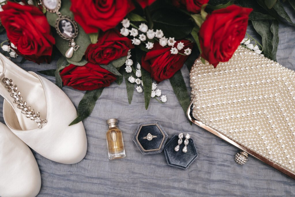 A flat lay of wedding accessories: satin shoes with jewelled straps, a pearl-embellished clutch, vintage jewellery, perfume, a ring in a box, pearl earrings, and a bouquet of red roses with white baby's breath on a soft grey fabric. © Aimee Lince Photography, Lincolnshire photographer