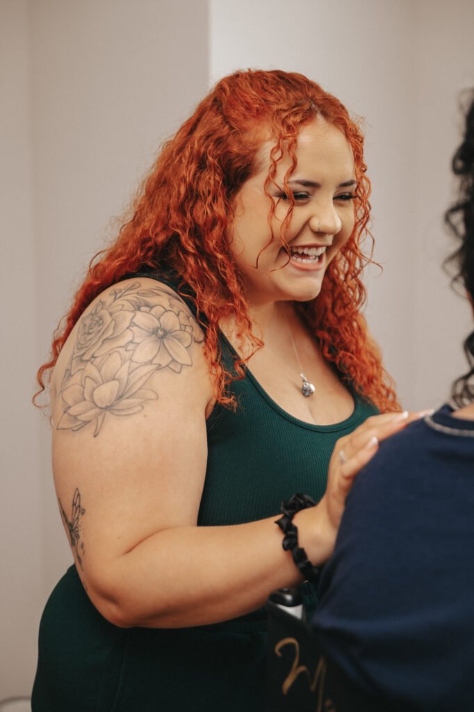 A woman with long curly red hair and a floral tattoo on her upper arm smiles warmly at another person. She wears a sleeveless dark green top and a necklace, and is inside a well-lit room, engaging in a friendly conversation. © Aimee Lince Photography, Lincolnshire photographer