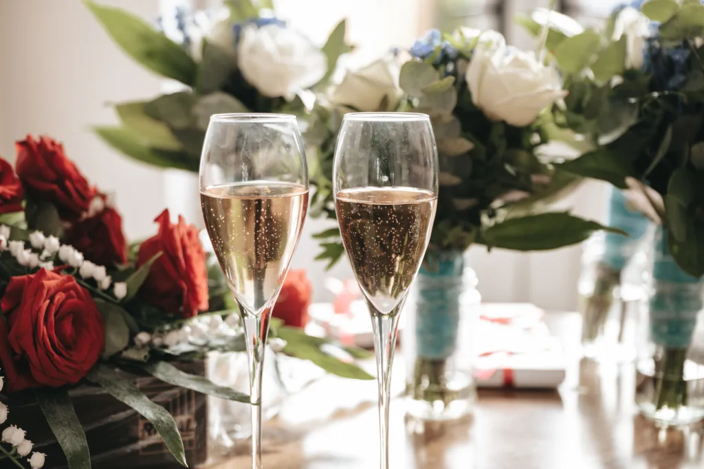 Two champagne flutes filled with sparkling wine sit on a table. In the background, there are bouquets of red roses and white flowers with green leaves, creating a festive and elegant atmosphere. © Aimee Lince Photography, Lincolnshire photographer