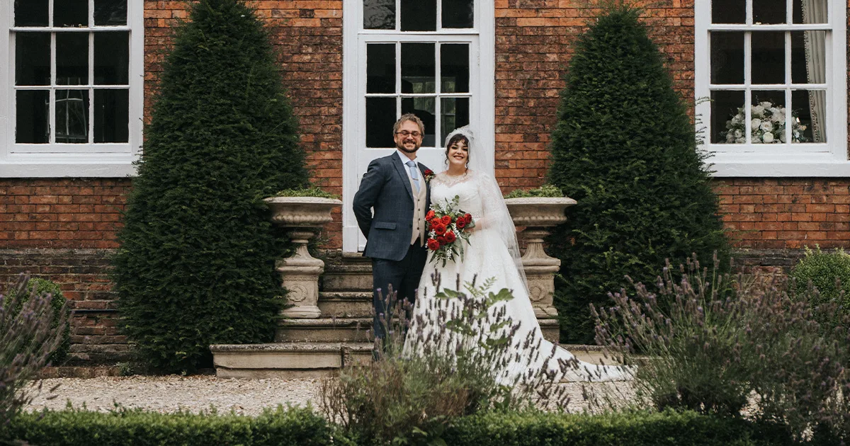 A bride and groom stand smiling together outdoors in front of a brick building with large windows and two tall, green bushes. The bride holds a red bouquet and wears a white dress and veil; the groom wears a suit and tie. © Aimee Lince Photography, wedding and lifestyle photographer in Lincolnshire