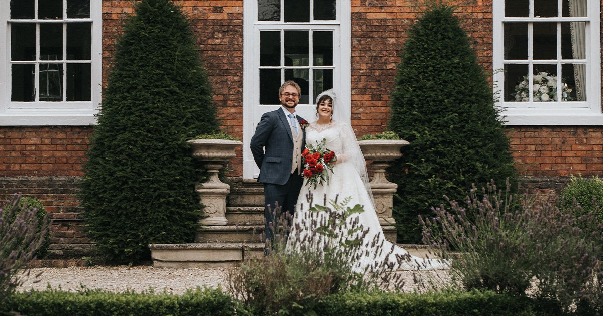 A bride and groom stand smiling together outdoors in front of a brick building with large windows and two tall, green bushes. The bride holds a red bouquet and wears a white dress and veil; the groom wears a suit and tie. © Aimee Lince Photography, wedding and lifestyle photographer in Lincolnshire