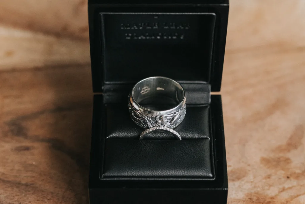 A silver ring with intricate detailing and a slim, curved band adorned with small stones, both beautifully displayed in an open black leather ring box on a wooden surface—perfectly captured by a wedding photographer for timeless memories. © Aimee Lince Photography, Lincolnshire photographer
