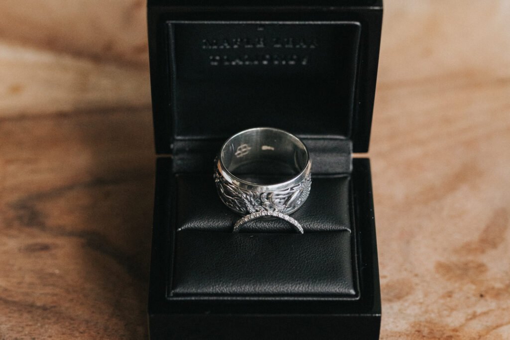 A silver ring with intricate detailing and a slim, curved band adorned with small stones, both beautifully displayed in an open black leather ring box on a wooden surface—perfectly captured by a wedding photographer for timeless memories. © Aimee Lince Photography, Lincolnshire photographer