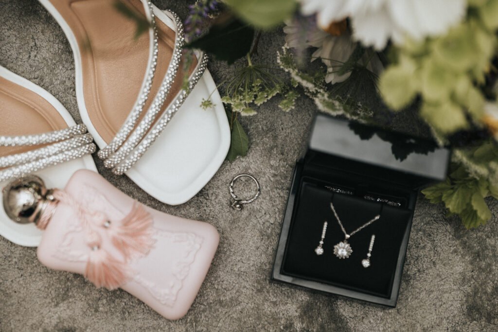 White sandals with sparkling straps, a pink perfume bottle, a diamond ring, and a black jewellery box displaying a matching necklace and earrings set are artfully arranged by the wedding photographer on a stone surface beside green and white flowers. © Aimee Lince Photography, Lincolnshire photographer