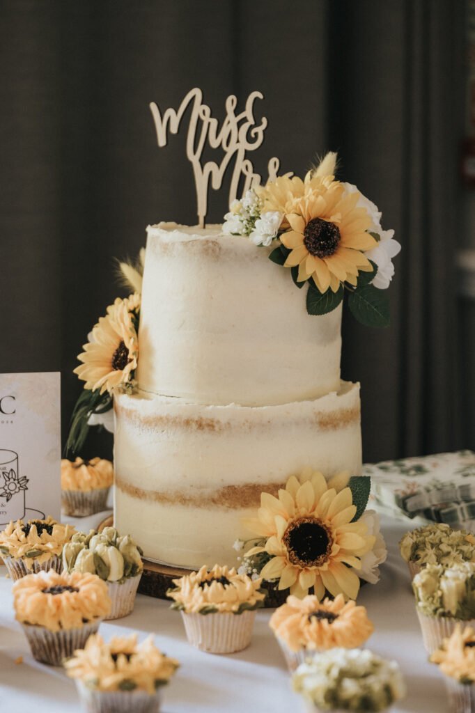 A two-tier semi-naked white wedding cake decorated with large artificial sunflowers and greenery sits on a table, captured beautifully by the wedding photographer. A wooden “Mrs & Mrs” topper crowns the cake, whilst matching cupcakes surround it. © Aimee Lince Photography, Lincolnshire photographer
