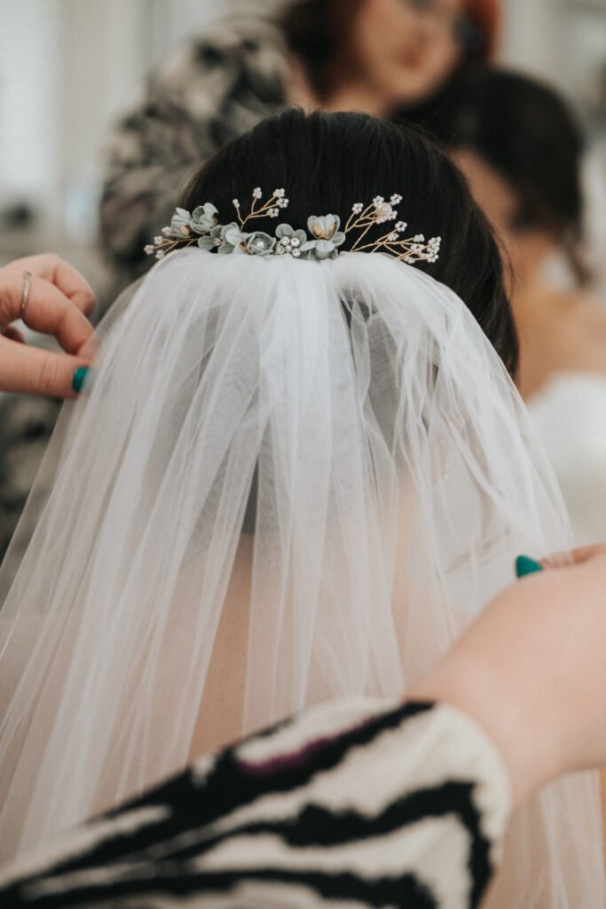 A bride’s hair is styled with a delicate white veil and a floral hairpiece of small white flowers and gold accents. A wedding photographer adjusts the veil, whilst another person, slightly blurred, appears in the background. © Aimee Lince Photography, Lincolnshire photographer