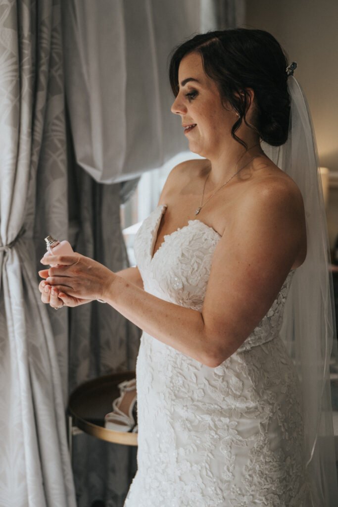 A bride in a white lace wedding dress and veil stands by a window, smiling as she holds a pink perfume bottle. Captured by a wedding photographer, her dark hair is styled up with loose curls, softly illuminated by natural light. Shoes rest on a small table behind her. © Aimee Lince Photography, Lincolnshire photographer