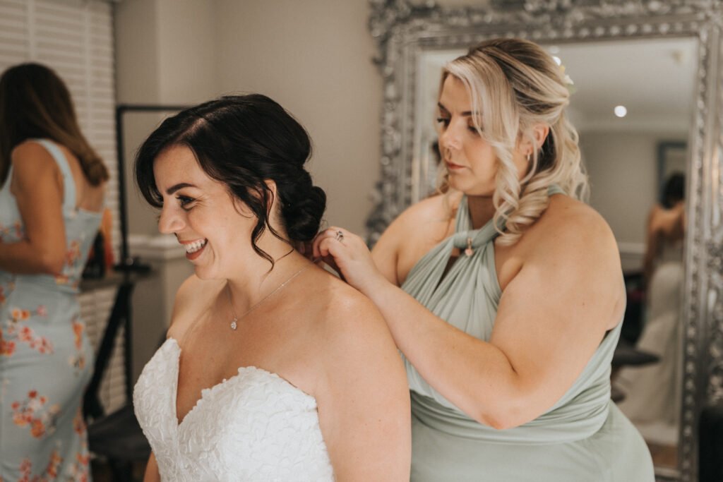 A smiling bride in a white lace strapless dress stands as a bridesmaid in sage green adjusts her necklace, captured perfectly by the wedding photographer. They are indoors with styled hair, an ornate mirror, and another woman in a floral dress in the background. © Aimee Lince Photography, Lincolnshire photographer