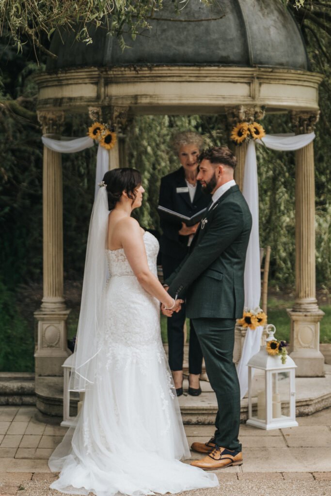 A bride and groom stand facing each other, holding hands, in front of a small gazebo decorated with white fabric and sunflowers. A photographer captures the moment as the officiant stands behind them, whilst the couple exchange vows outdoors. © Aimee Lince Photography, Lincolnshire photographer
