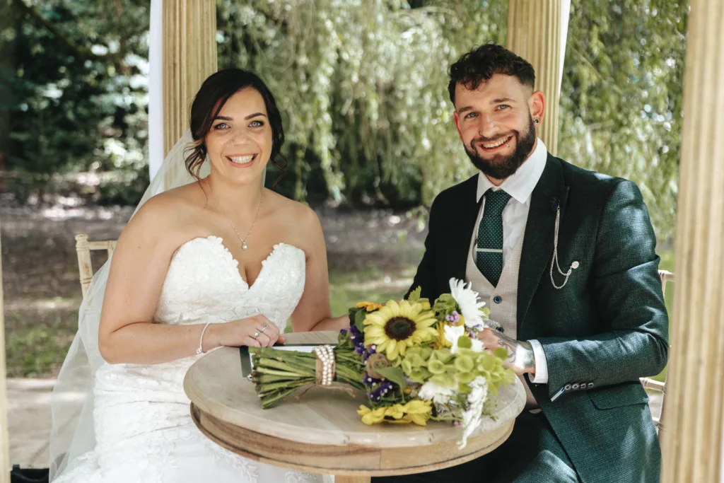 A smiling bride in a strapless white gown and a groom in a dark suit sit at a round wooden table outdoors, under leafy trees. Captured by their wedding photographer, they celebrate with sunflowers and green flowers arranged on the table. © Aimee Lince Photography, Lincolnshire photographer