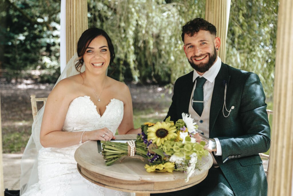 A smiling bride in a strapless white gown and a groom in a dark suit sit at a round wooden table outdoors, under leafy trees. Captured by their wedding photographer, they celebrate with sunflowers and green flowers arranged on the table. © Aimee Lince Photography, Lincolnshire photographer