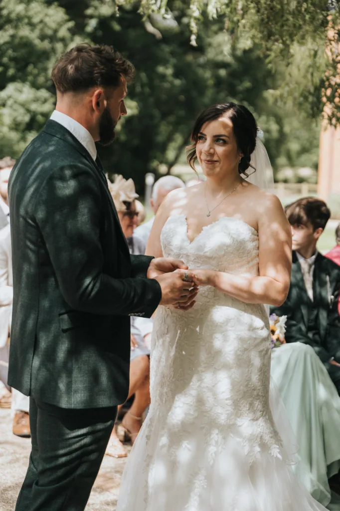 A groom in a dark suit holds the hand of a bride in a white strapless dress during an outdoor wedding ceremony, as their wedding photographer captures the moment with guests watching under dappled sunlight and leafy trees. © Aimee Lince Photography, Lincolnshire photographer