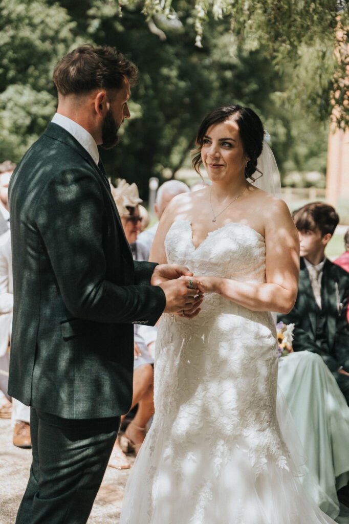 A groom in a dark suit holds the hand of a bride in a white strapless dress during an outdoor wedding ceremony, as their wedding photographer captures the moment with guests watching under dappled sunlight and leafy trees. © Aimee Lince Photography, Lincolnshire photographer
