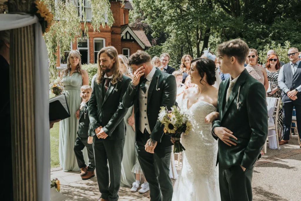 A groom wipes tears from his face during an outdoor wedding ceremony, captured by the wedding photographer. The bride in a white dress holds a bouquet as ushers and bridesmaids in green and guests watch this emotional garden moment. © Aimee Lince Photography, Lincolnshire photographer