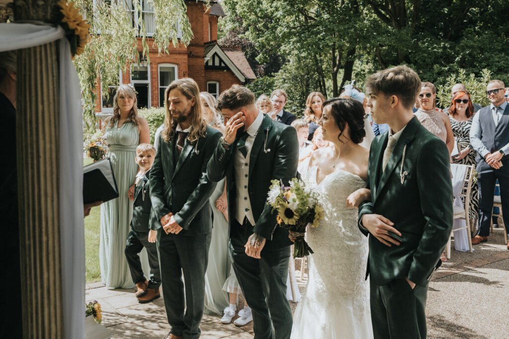 A groom wipes tears from his face during an outdoor wedding ceremony, captured by the wedding photographer. The bride in a white dress holds a bouquet as ushers and bridesmaids in green and guests watch this emotional garden moment. © Aimee Lince Photography, Lincolnshire photographer