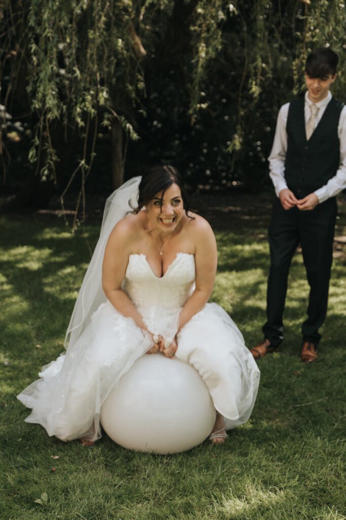 A bride in a white wedding dress and veil smiles whilst sitting on a large exercise ball on grass. A photographer captures the playful moment as her groom stands behind her, both smiling under the shade of trees. The mood is joyful and fun. © Aimee Lince Photography, Lincolnshire photographer