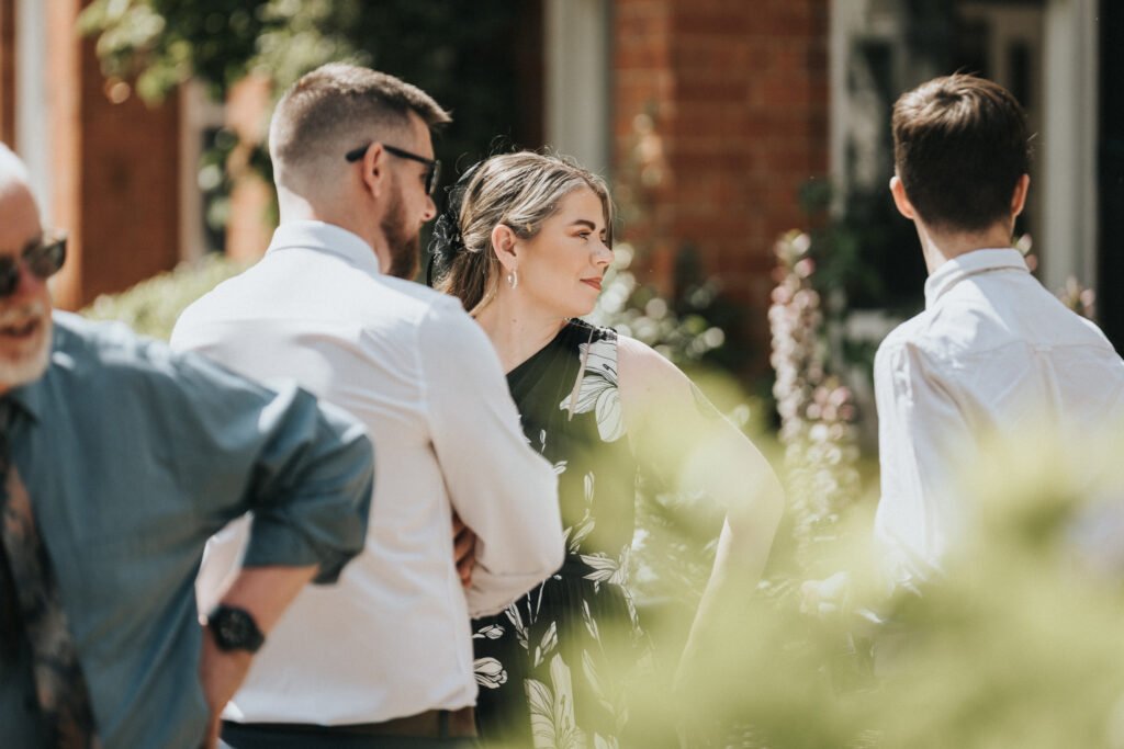 Four adults stand outside in sunlight, engaged in conversation. A wedding photographer captures a woman in a black and white dress talking to a man in a white shirt, while another man in sunglasses and a blue shirt stands nearby. Brick building and greenery form the backdrop. © Aimee Lince Photography, Lincolnshire photographer