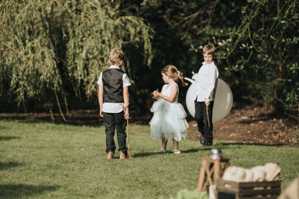 Three children stand on a grassy lawn; two boys in dress shirts and trousers, one holding a large white balloon, and a girl in a white tulle dress. Sunlight filters through trees as a wedding photographer captures this warm, playful outdoor scene. © Aimee Lince Photography, Lincolnshire photographer