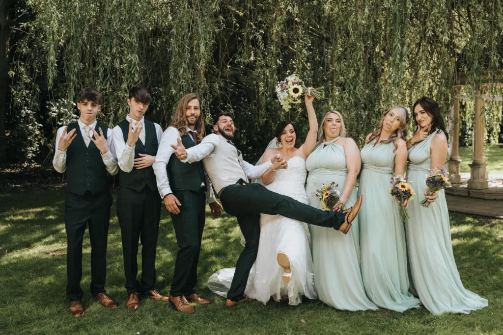 A wedding photographer captures a joyful outdoor party under a tree. The bride in white lifts her bouquet beside the groom, playfully kicking his foot, as bridesmaids in mint and groomsmen in dark suits strike expressive poses. © Aimee Lince Photography, Lincolnshire photographer