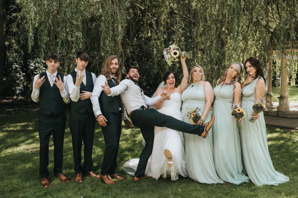 A wedding photographer captures a joyful outdoor party under a tree. The bride in white lifts her bouquet beside the groom, playfully kicking his foot, as bridesmaids in mint and groomsmen in dark suits strike expressive poses. © Aimee Lince Photography, Lincolnshire photographer