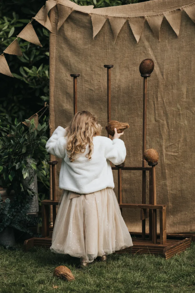A young girl in a white jacket and tulle skirt stands outdoors, facing a coconut shy game captured by a wedding photographer. She holds a coconut, ready to throw at wooden targets beneath hessian bunting, with lush greenery completing the rustic scene. © Aimee Lince Photography, Lincolnshire photographer
