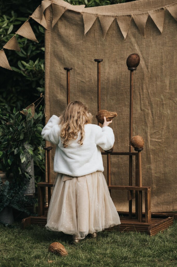 A young girl in a white jacket and tulle skirt stands outdoors, facing a coconut shy game captured by a wedding photographer. She holds a coconut, ready to throw at wooden targets beneath hessian bunting, with lush greenery completing the rustic scene. © Aimee Lince Photography, Lincolnshire photographer