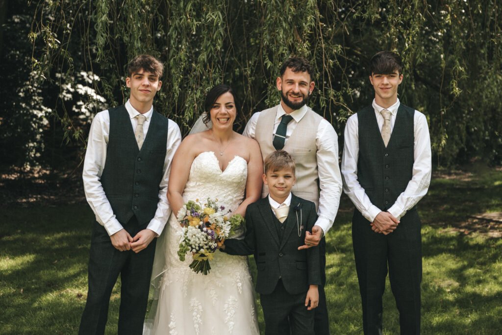 A bride in a strapless white gown and groom in a waistcoat and tie stand outdoors with three boys in matching waistcoats and ties. Captured by a wedding photographer, they smile under hanging greenery as sunlight filters through the trees. © Aimee Lince Photography, Lincolnshire photographer