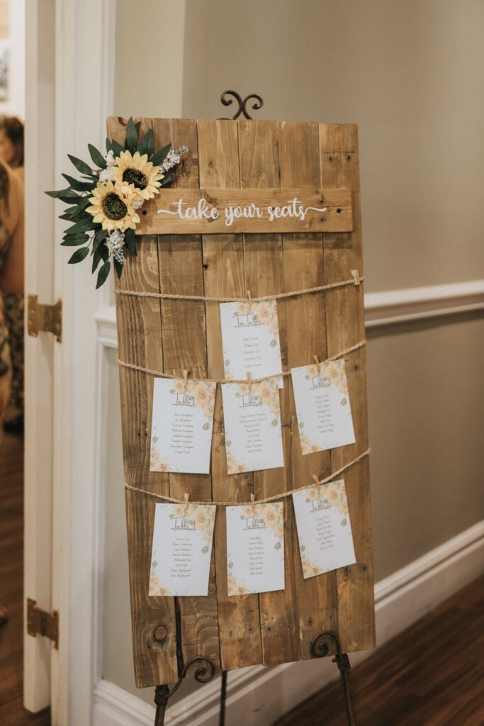 A rustic wooden seating chart decorated with sunflowers and greenery is displayed on an easel—perfect for a wedding photographer to capture. "Take your seats" tops the printed cards with guest names, hanging from twine rows by pegs. © Aimee Lince Photography, Lincolnshire photographer