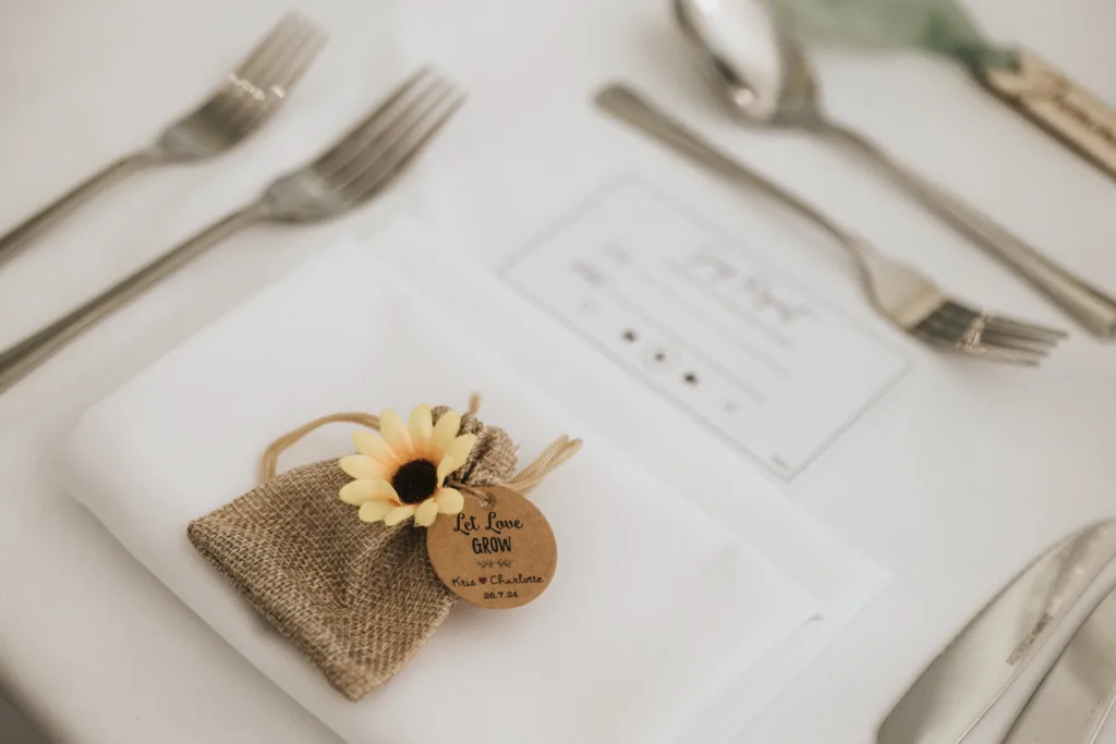 A place setting with a white serviette, cutlery, and a hessian pouch topped with a yellow artificial sunflower awaits the wedding photographer’s keen eye. The pouch’s wooden tag reads “Let Love Grow” with “Alex & Charlotte, 27.7.24.”. © Aimee Lince Photography, Lincolnshire photographer