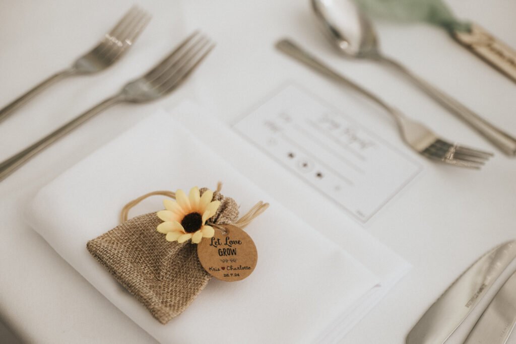 A place setting with a white serviette, cutlery, and a hessian pouch topped with a yellow artificial sunflower awaits the wedding photographer’s keen eye. The pouch’s wooden tag reads “Let Love Grow” with “Alex & Charlotte, 27.7.24.”. © Aimee Lince Photography, Lincolnshire photographer
