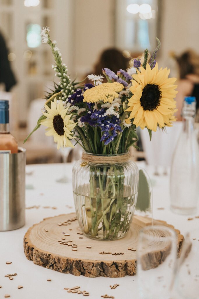 A glass jar filled with sunflowers and wildflowers sits on a wooden slab centrepiece on a white tablecloth, perfectly styled for a wedding photographer. Bottles, glasses, and wooden confetti surround the arrangement in a softly lit, blurred background. © Aimee Lince Photography, Lincolnshire photographer