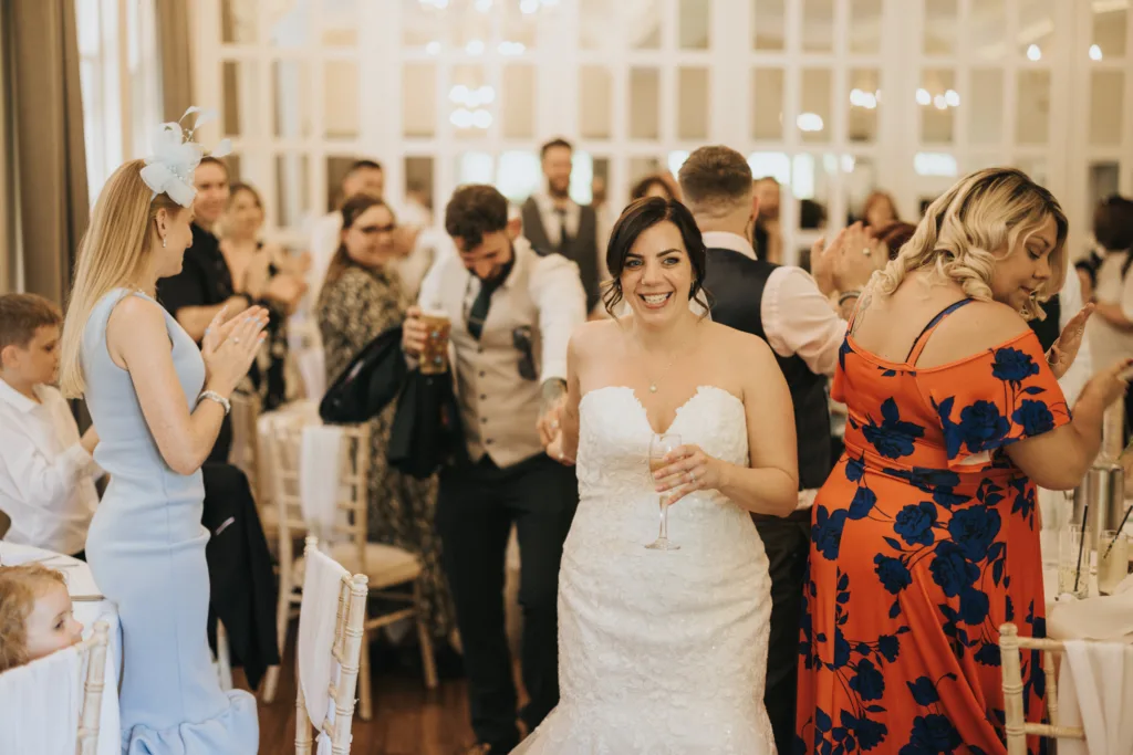 A bride in a strapless white gown smiles whilst holding a champagne flute at a wedding reception, beautifully captured by a skilled wedding photographer. Guests in colourful outfits clap and celebrate around her in an elegant, chandelier-lit room. © Aimee Lince Photography, Lincolnshire photographer