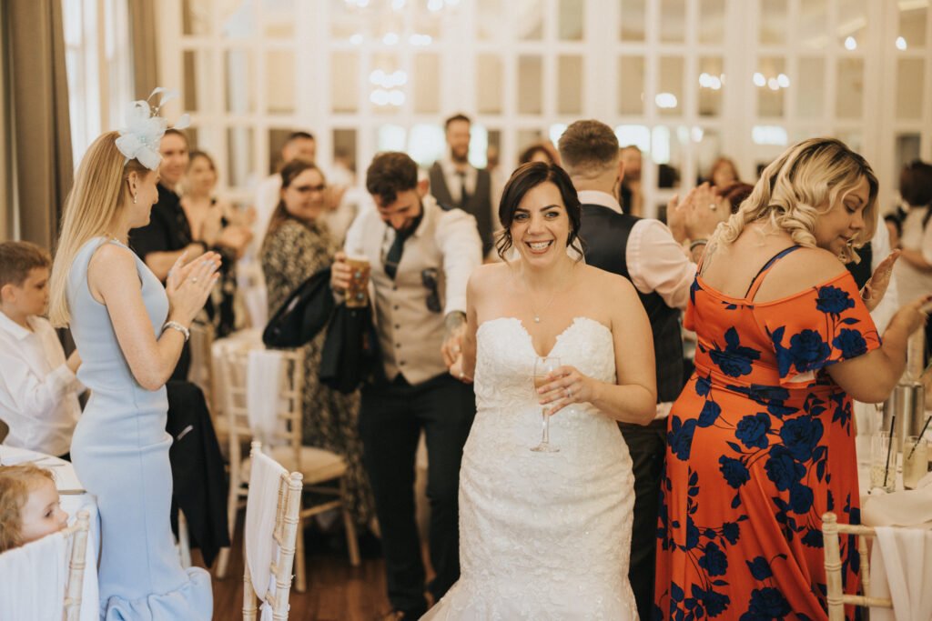 A bride in a strapless white gown smiles whilst holding a champagne flute at a wedding reception, beautifully captured by a skilled wedding photographer. Guests in colourful outfits clap and celebrate around her in an elegant, chandelier-lit room. © Aimee Lince Photography, Lincolnshire photographer