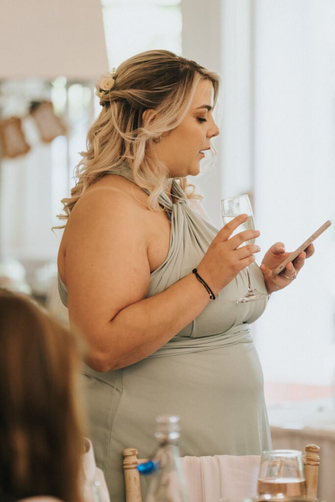 A woman in a pale green dress holds a wine glass and reads from her mobile, giving a speech at a formal event. A wedding photographer captures her long, wavy blonde hair with a small flower delicately pinned in the back. © Aimee Lince Photography, Lincolnshire photographer