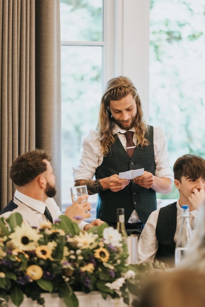 A man with long hair and a beard stands reading from a piece of paper at a table decorated with flowers, as a wedding photographer captures the moment. Two men in waistcoats and ties listen attentively; one raises a glass amid sunlight and grey curtains. © Aimee Lince Photography, Lincolnshire photographer