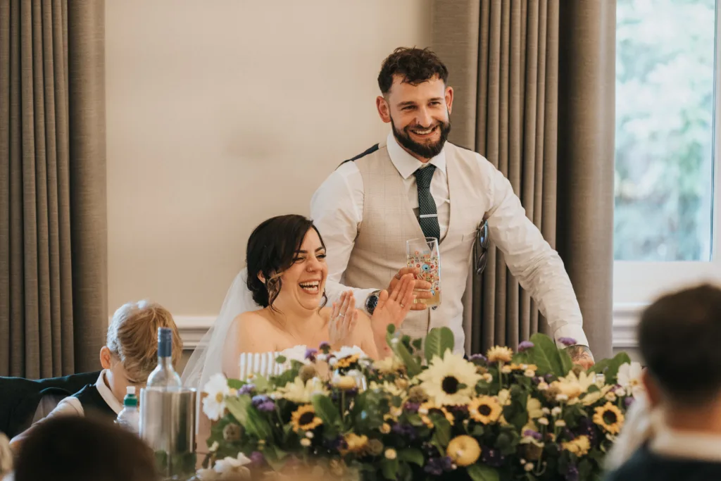 A groom stands smiling behind a seated bride, who is laughing and clapping. Captured by a wedding photographer, they sit at a table decorated with sunflowers and purple blooms. The groom wears a waistcoat and tie; the bride, a white dress and veil. Light streams through the window. © Aimee Lince Photography, Lincolnshire photographer