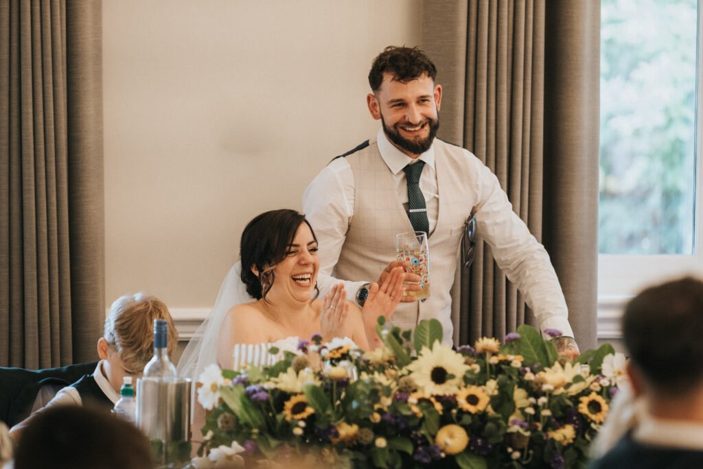 A groom stands smiling behind a seated bride, who is laughing and clapping. Captured by a wedding photographer, they sit at a table decorated with sunflowers and purple blooms. The groom wears a waistcoat and tie; the bride, a white dress and veil. Light streams through the window. © Aimee Lince Photography, Lincolnshire photographer