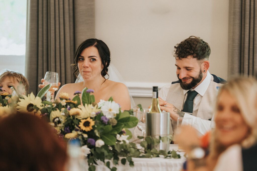 A bride in a strapless dress holds a glass and smiles with a surprised expression beside a bearded groom, both seated at a table decorated with sunflowers and greenery, as the wedding photographer captures the joyful moment amongst guests. © Aimee Lince Photography, Lincolnshire photographer